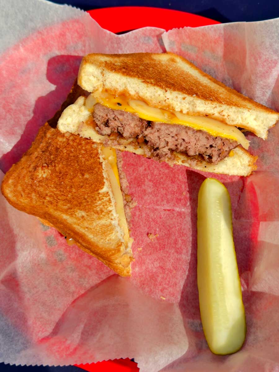 Grilled cheese burger sandwich halved on red tray with a pickle spear from Fat Daddy’s Menu.